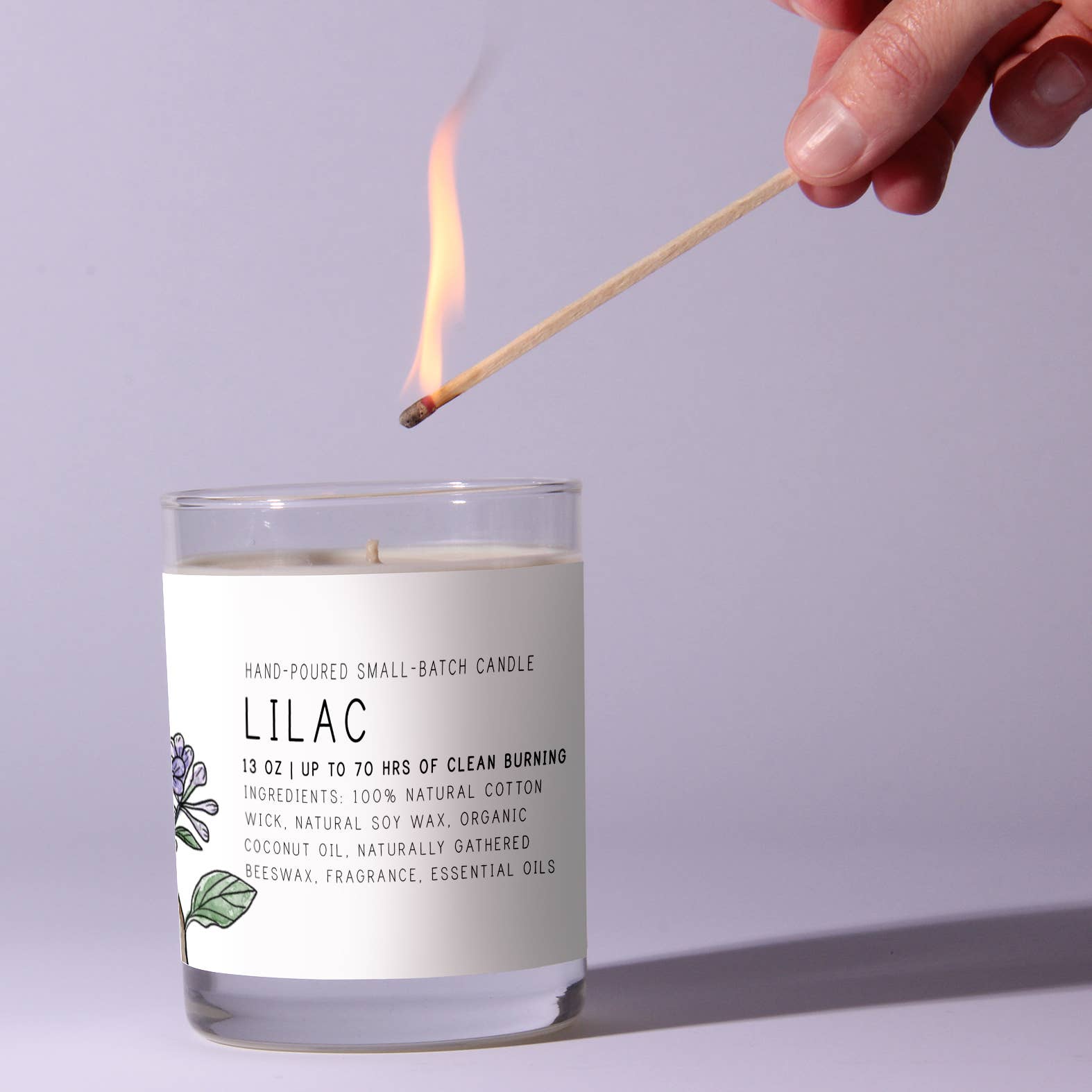Candle labeled 'Lilac' being lit with a match against a light purple background