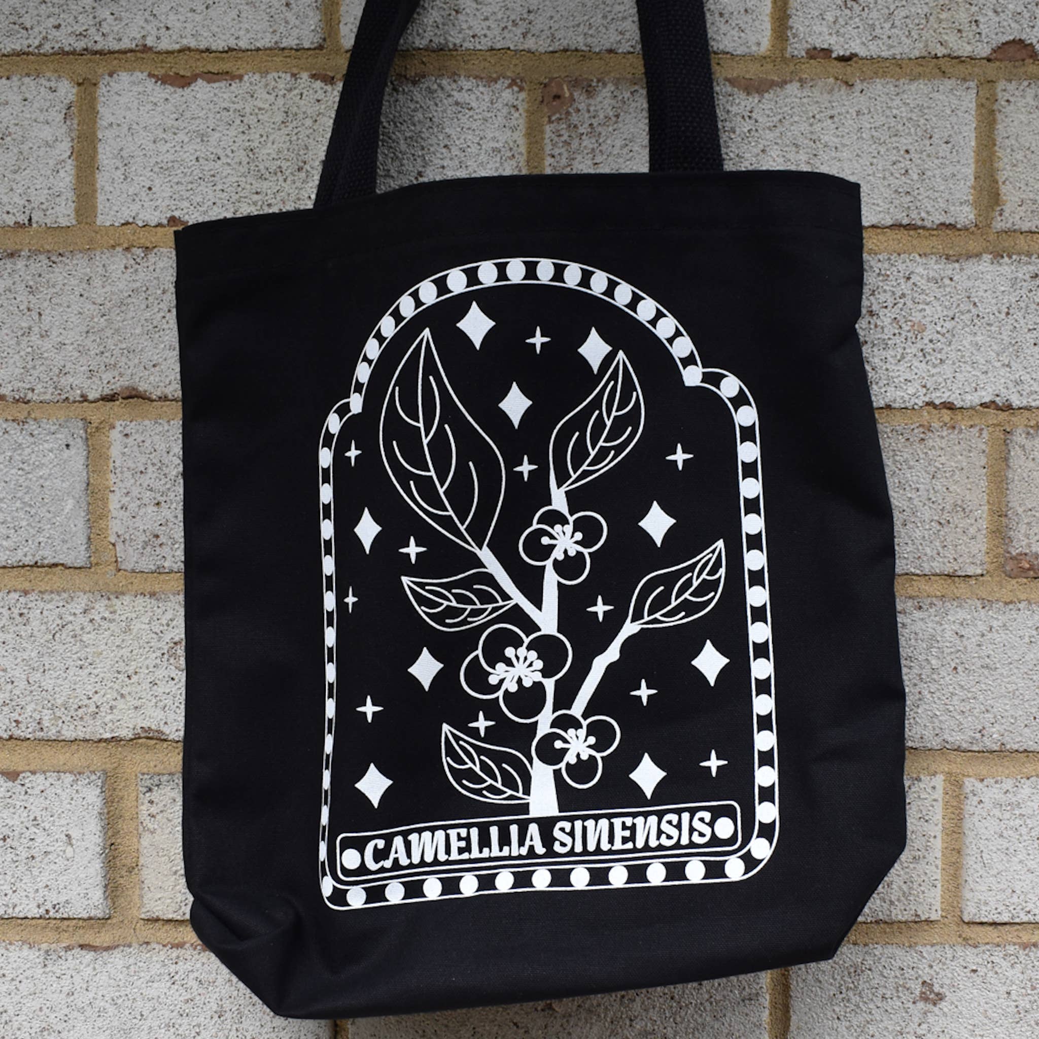 Black tote bag with white floral design and 'Camellia Sinensis' text against a brick wall.