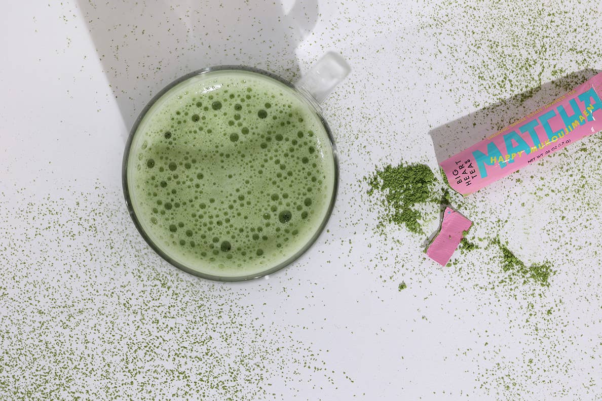 Matcha green tea powder with a glass of matcha latte on a white surface.