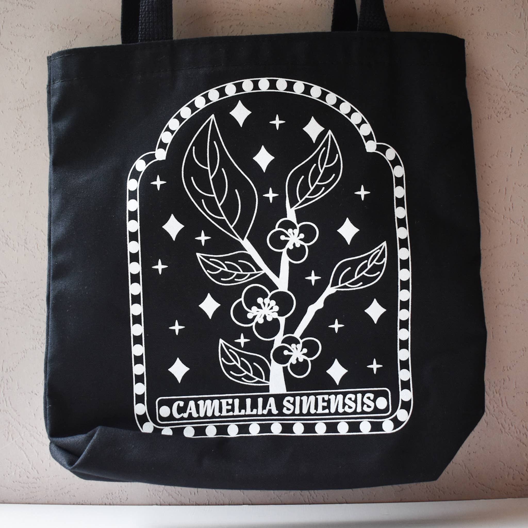 Black tote bag with white floral design and 'Camellia Sinensis' text on a neutral background