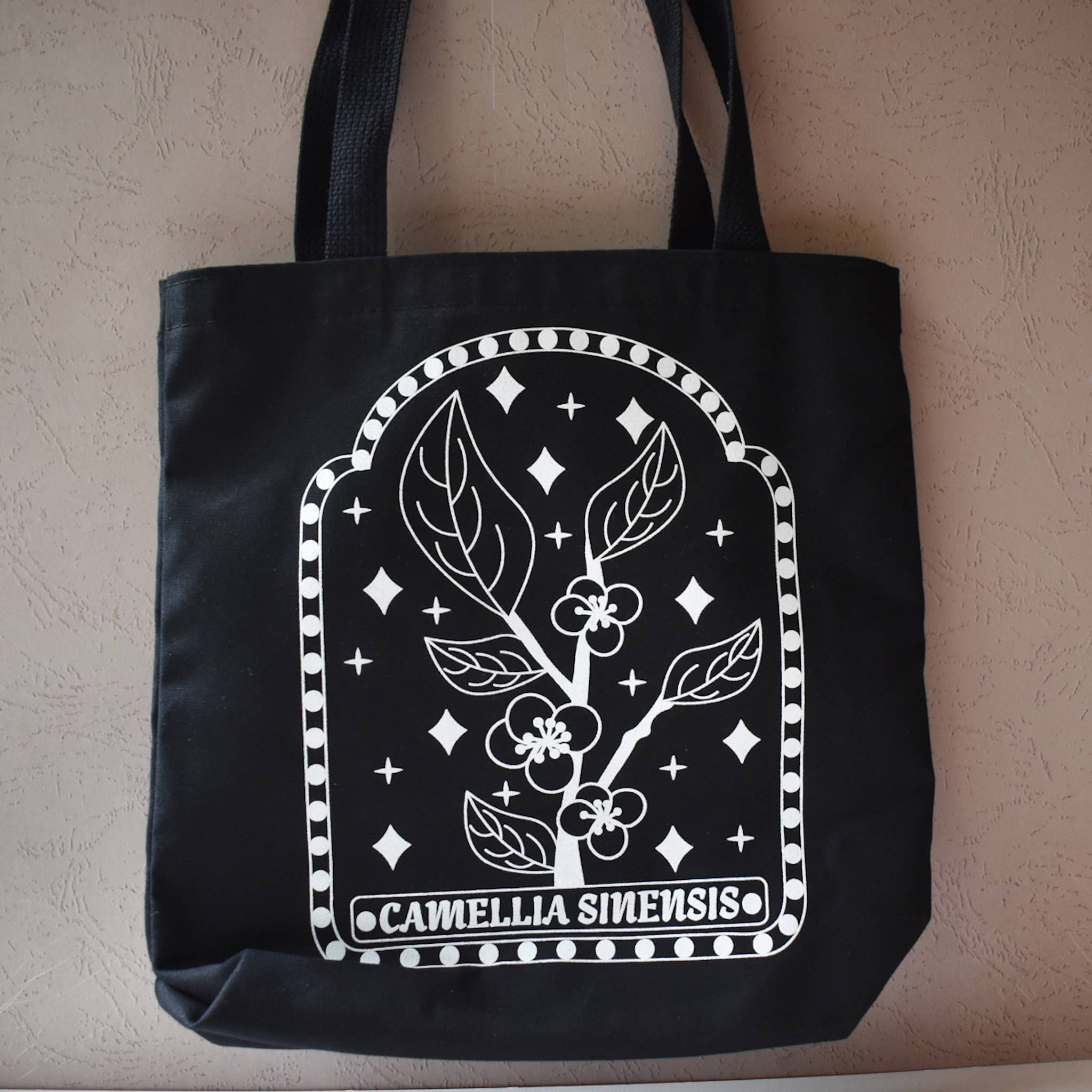 Black tote bag with white floral design and 'Camellia Sinensis' text on a beige background