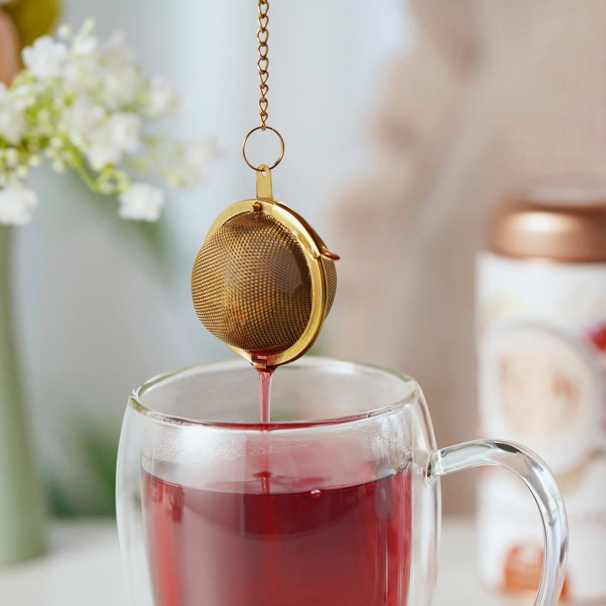 Gold tea infuser being used to steep tea in a glass mug with a blurred background.