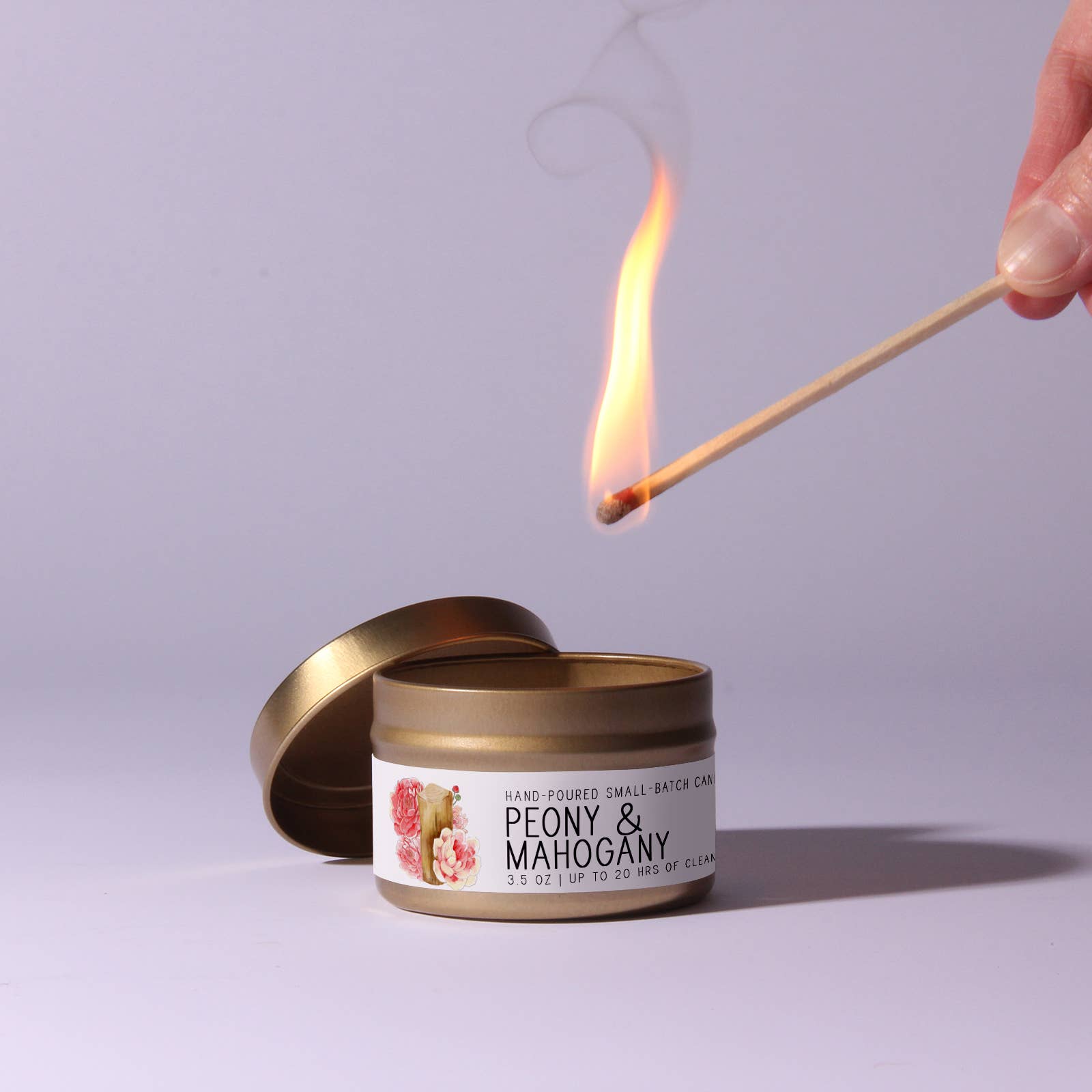 Candle labeled 'Peony & Mahogany' being lit with a match against a light purple background