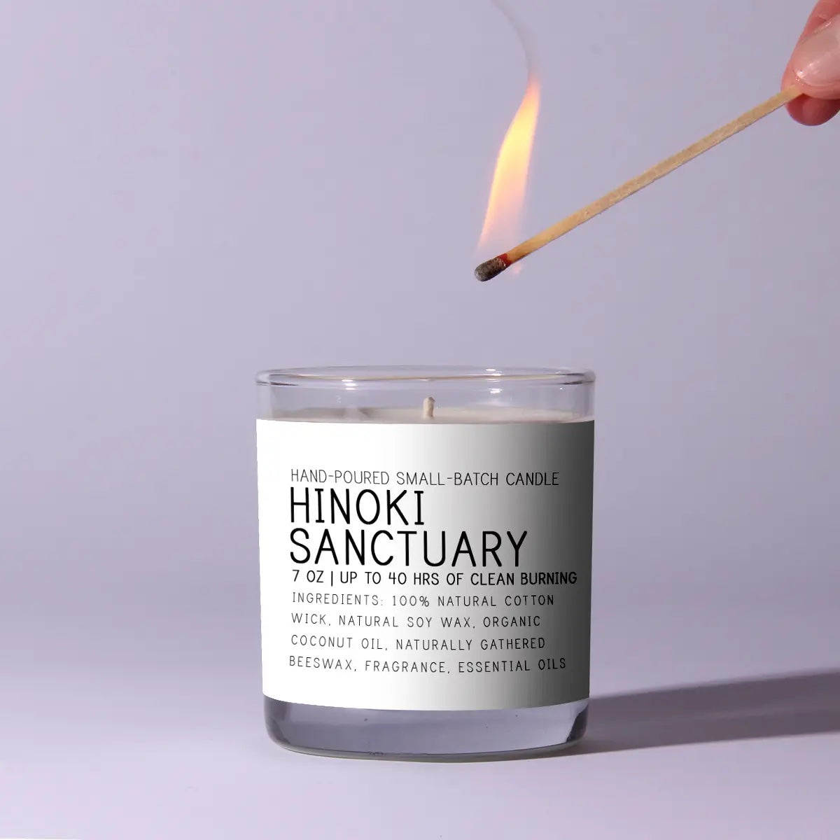 Candle labeled 'Hinoki Sanctuary' being lit with a match against a light purple background