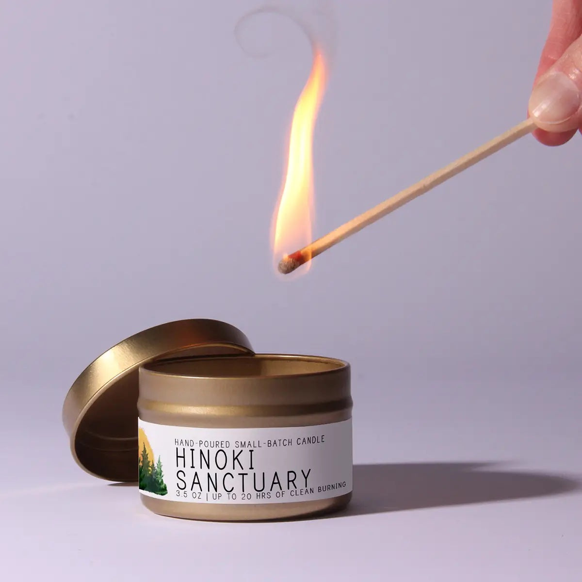 Hinoki Sanctuary candle being lit with a match on a light purple background