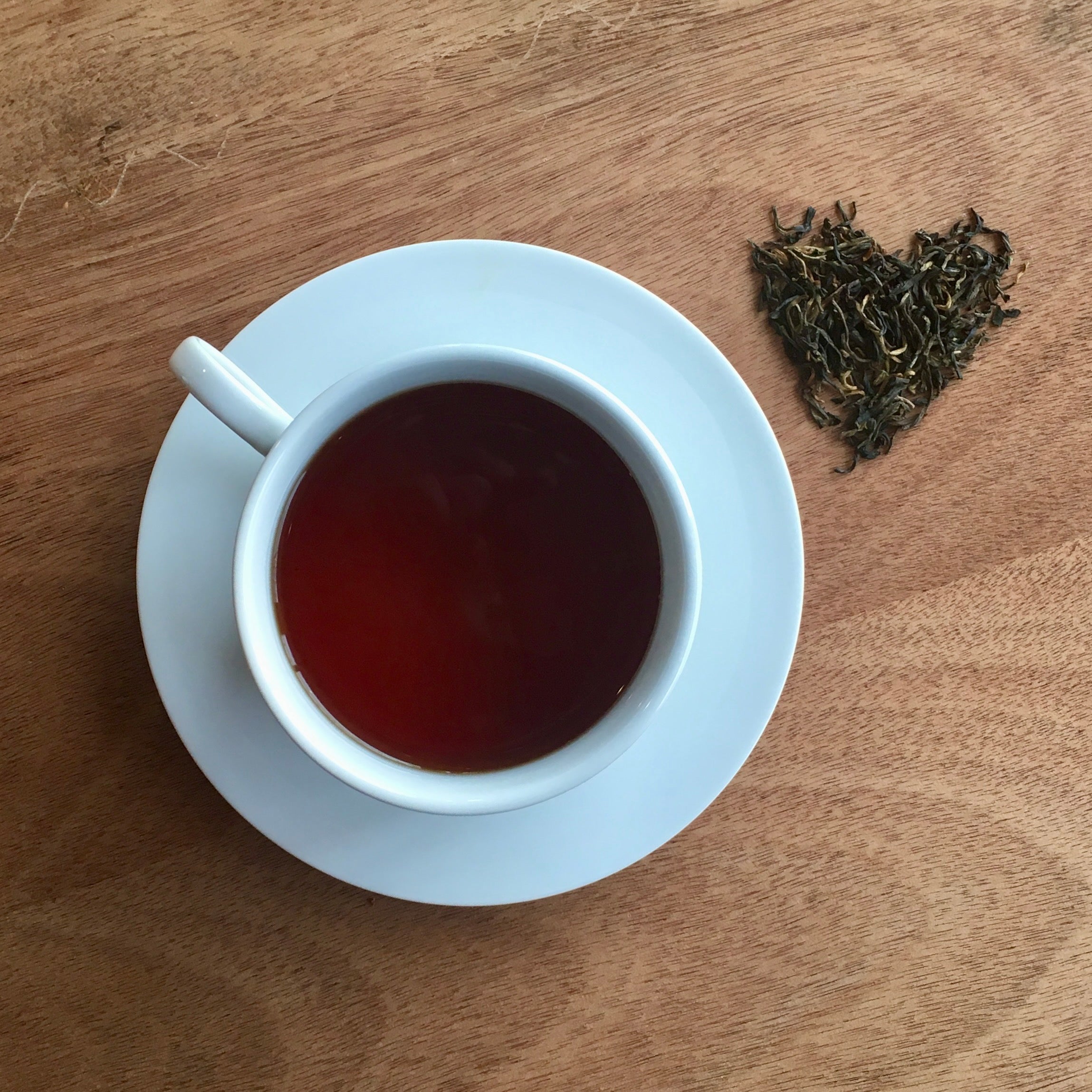 White cup of tea on a wooden surface with dried tea leaves forming a heart shape.