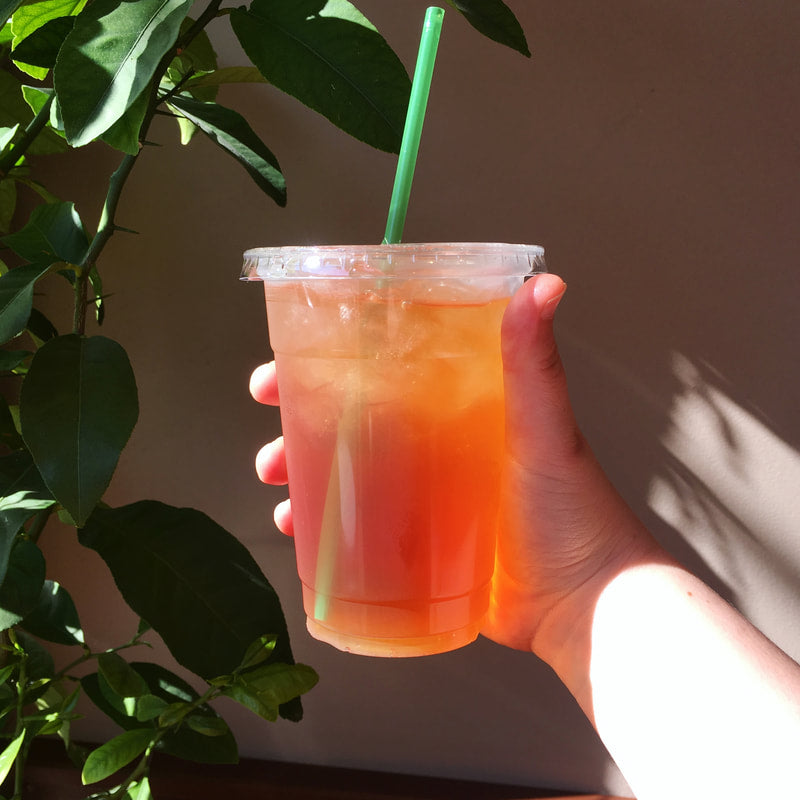 Hand holding a plastic cup with iced tea and a green straw, with a plant in the background.
