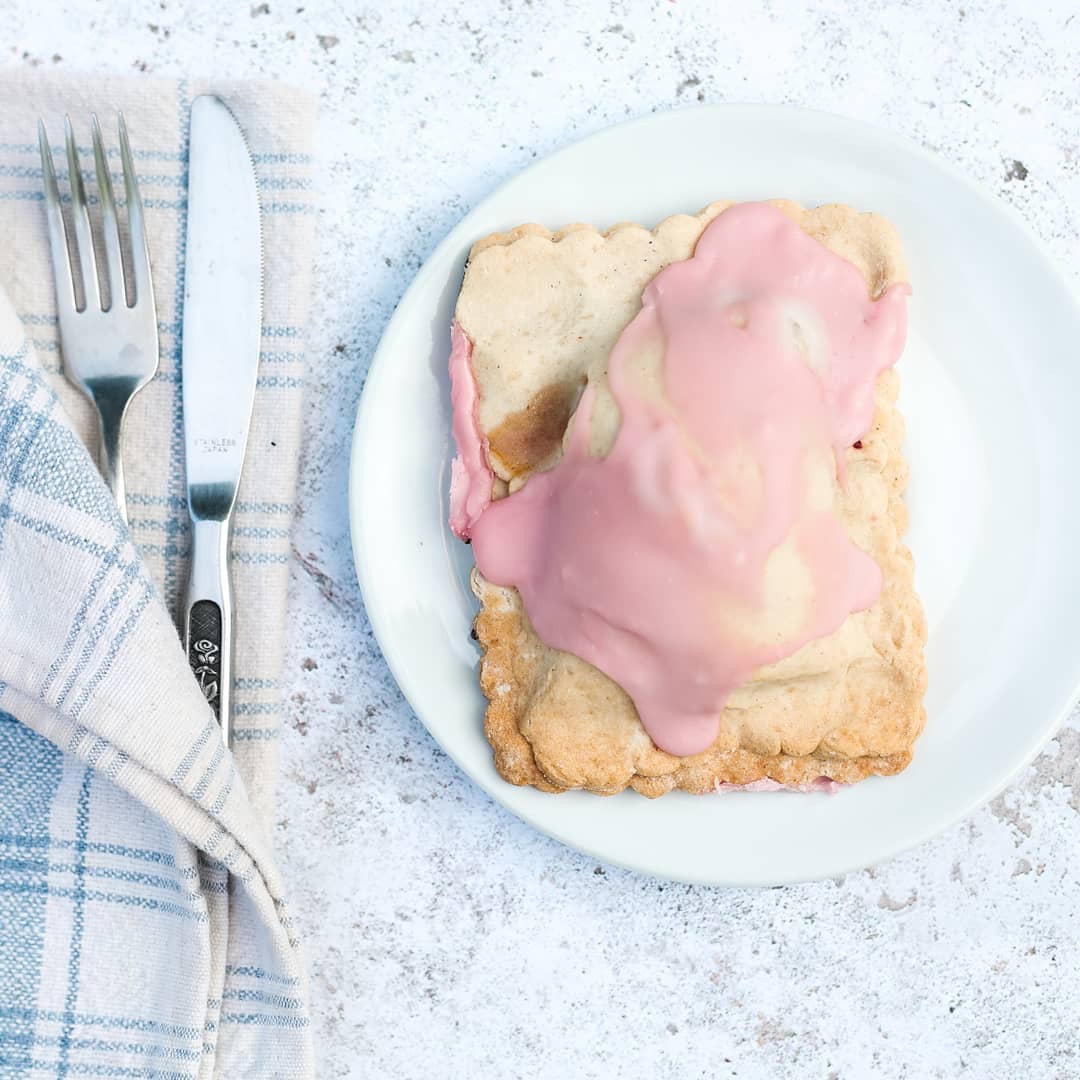 Pastry with pink glaze on a white plate with a fork and knife, on a textured surface.