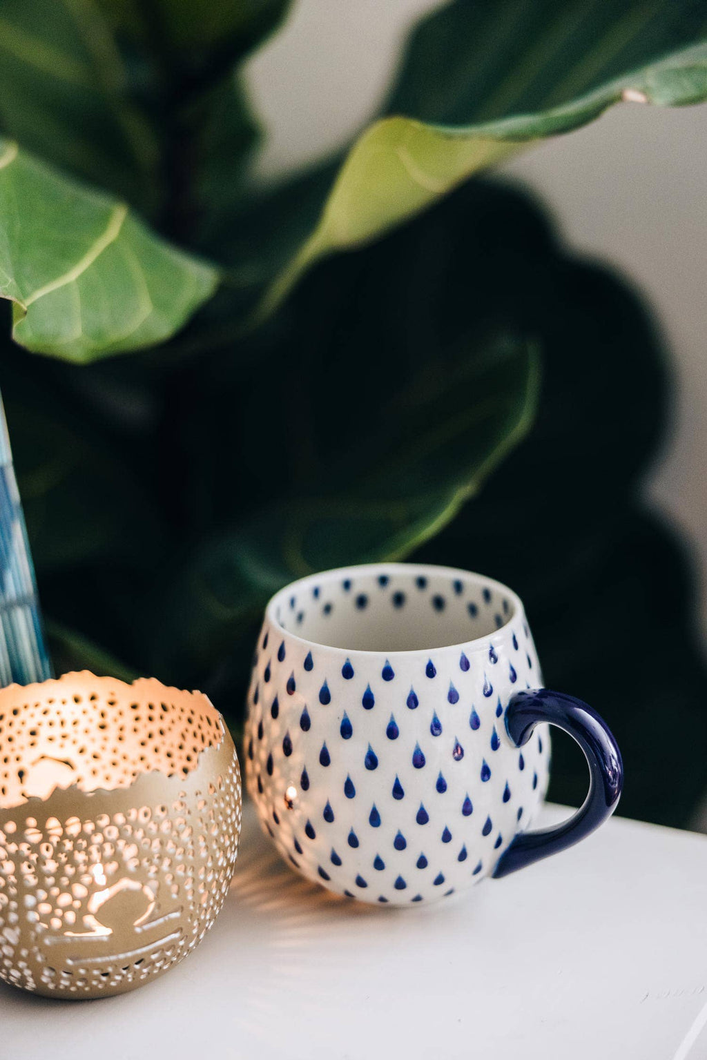 White mug with blue pattern and handle next to a lit candle on a surface with green foliage in the background