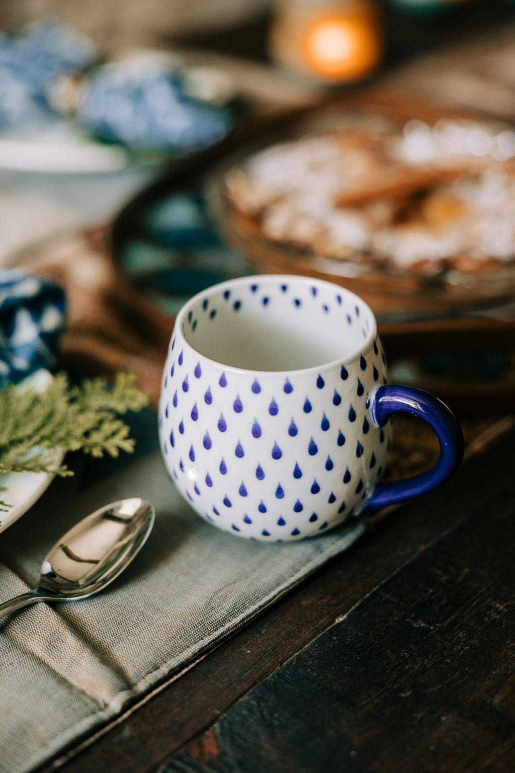 White mug with blue polka dot pattern and blue handle on a wooden surface with a spoon and blurred background