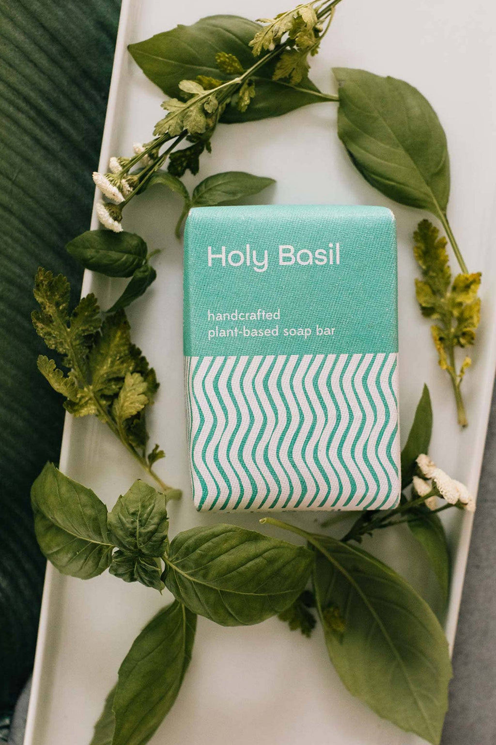 Handcrafted plant-based soap bar with 'Holy Basil' packaging on a white plate surrounded by basil leaves.