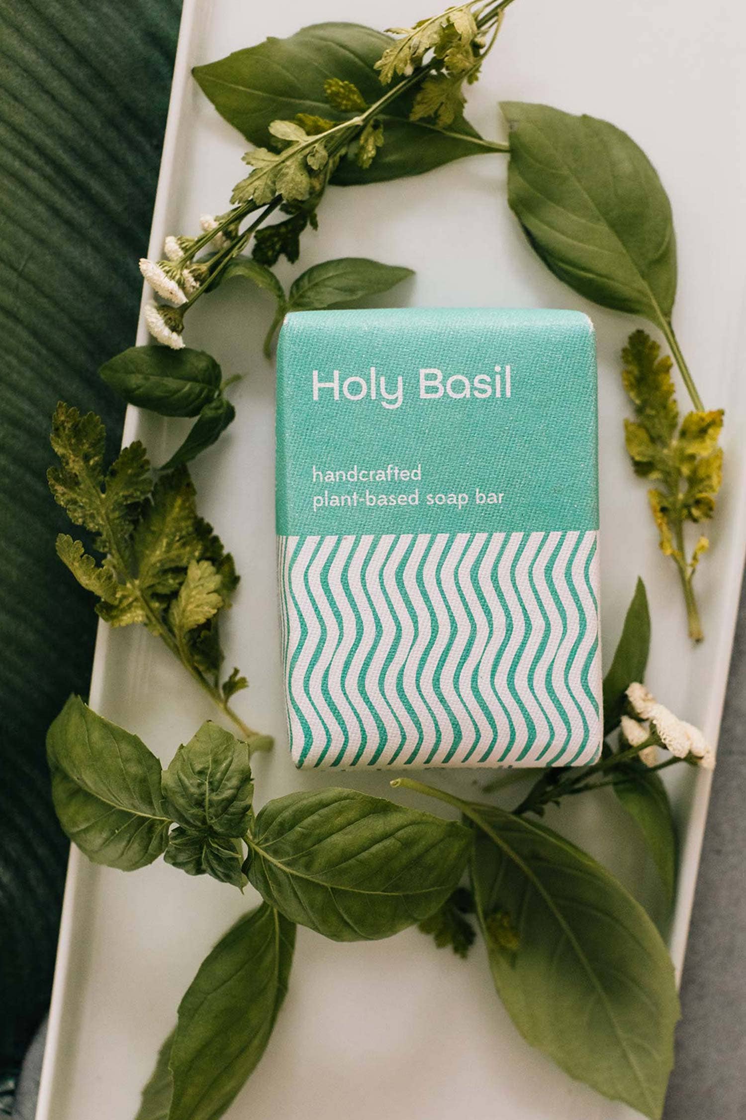 Handcrafted plant-based soap bar with 'Holy Basil' packaging on a white plate surrounded by basil leaves.