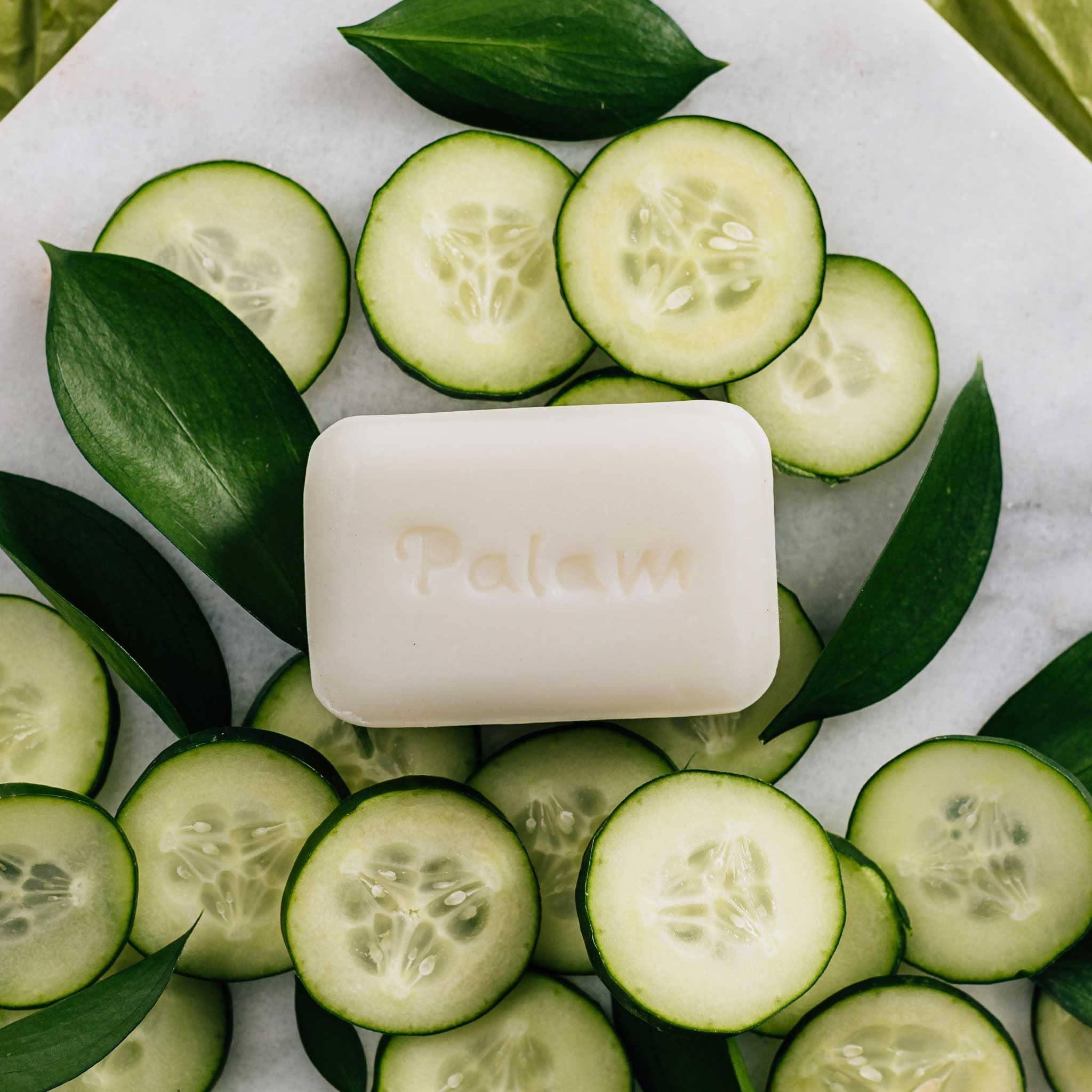 Bar of soap surrounded by sliced cucumbers and leaves on a marble surface