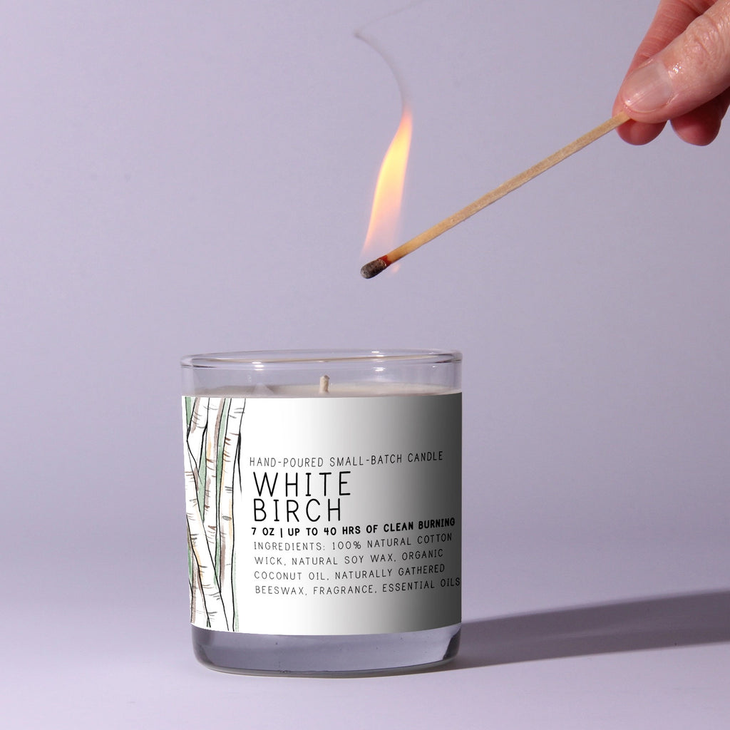 Candle labeled 'White Birch' being lit with a match against a light purple background