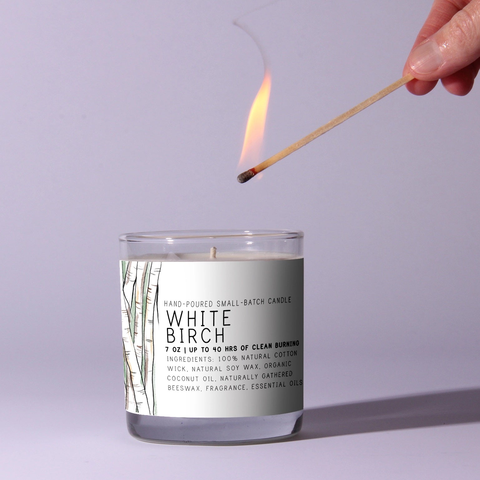 Candle labeled 'White Birch' being lit with a match against a light purple background