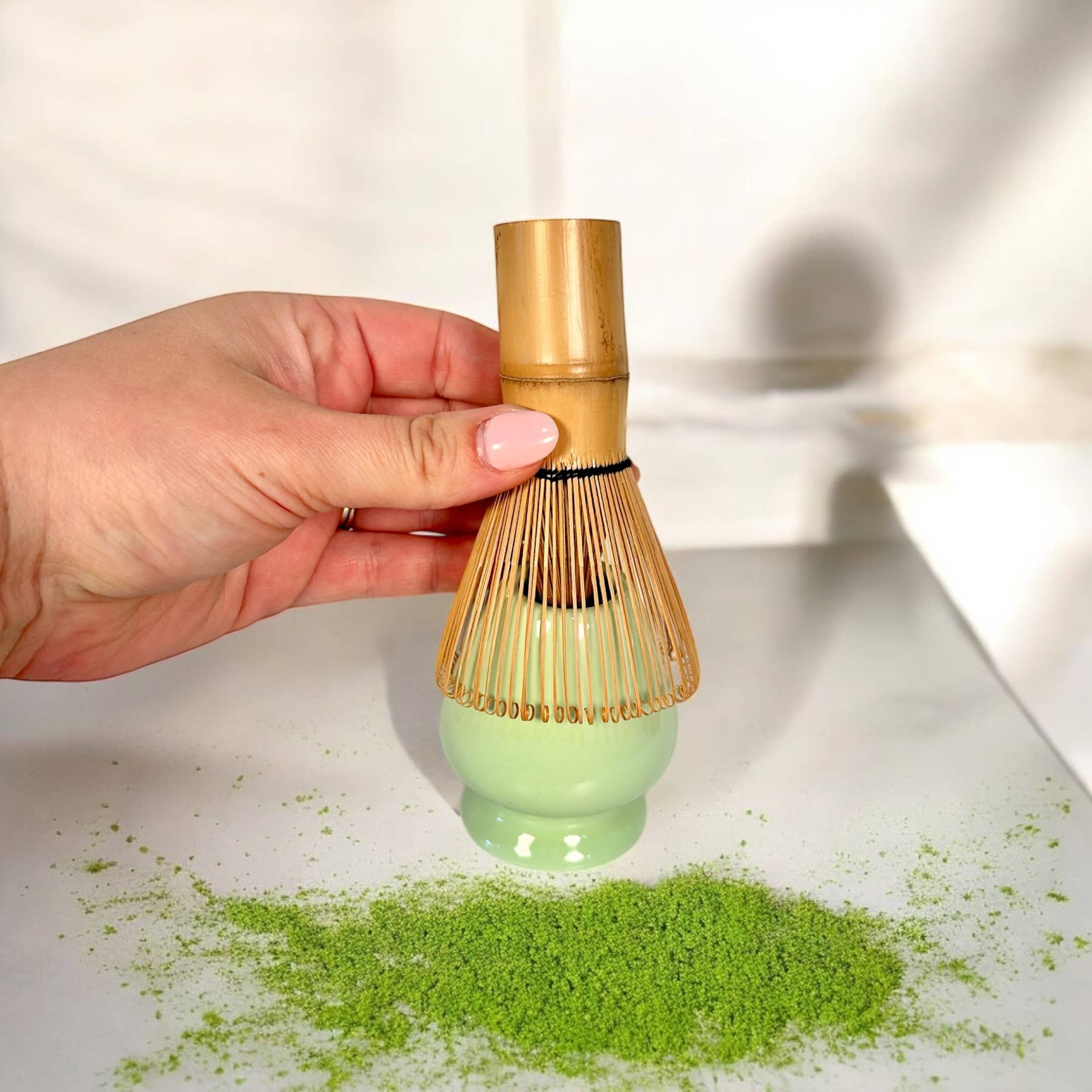 Hand holding a bamboo whisk over matcha green tea powder on a white surface