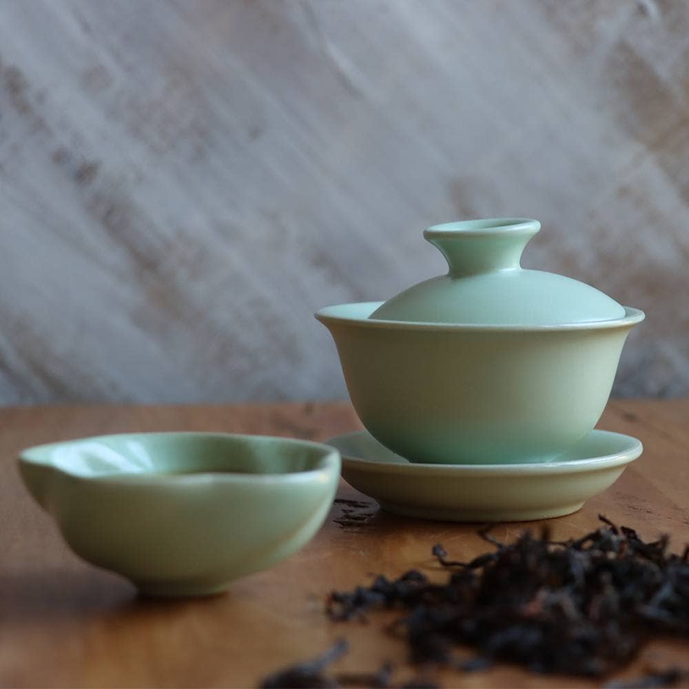 Green ceramic gaiwan on a wooden surface with tea leaves.