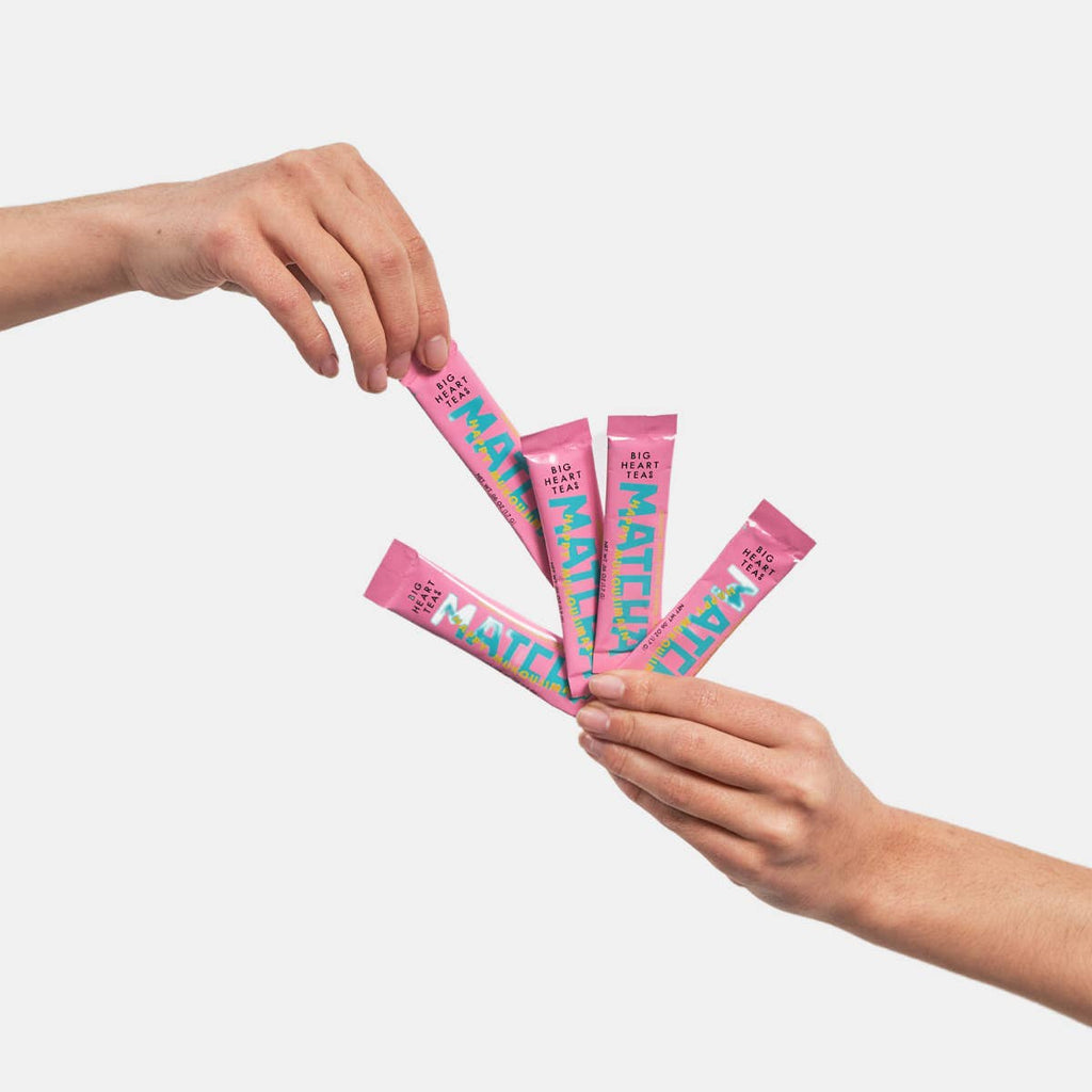 Two hands holding pink packets of matcha with colorful text on a light gray background