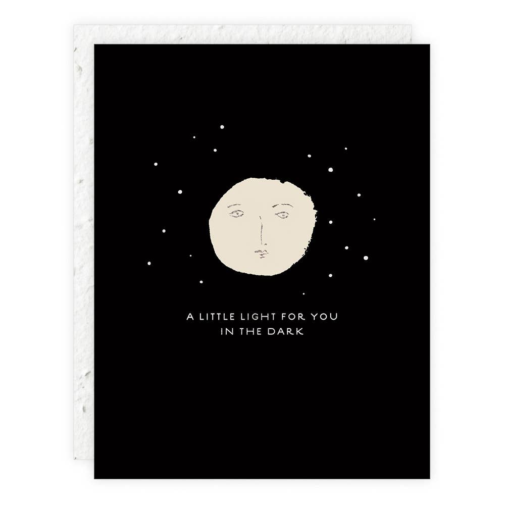 Black card with a beige moon face and text 'A little light for you in the dark' on a white background