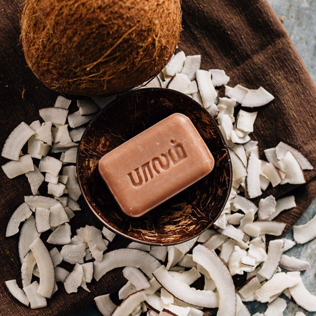 Coconut bar soap in a coconut shell surrounded by coconut pieces on a wooden surface.