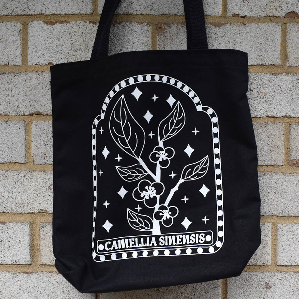 Black tote bag with white floral design and 'Camellia Sinensis' text against a brick wall.