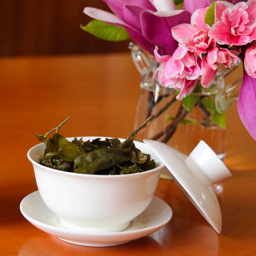 White teacup with green tea leaves on a wooden surface, with pink flowers in the background.