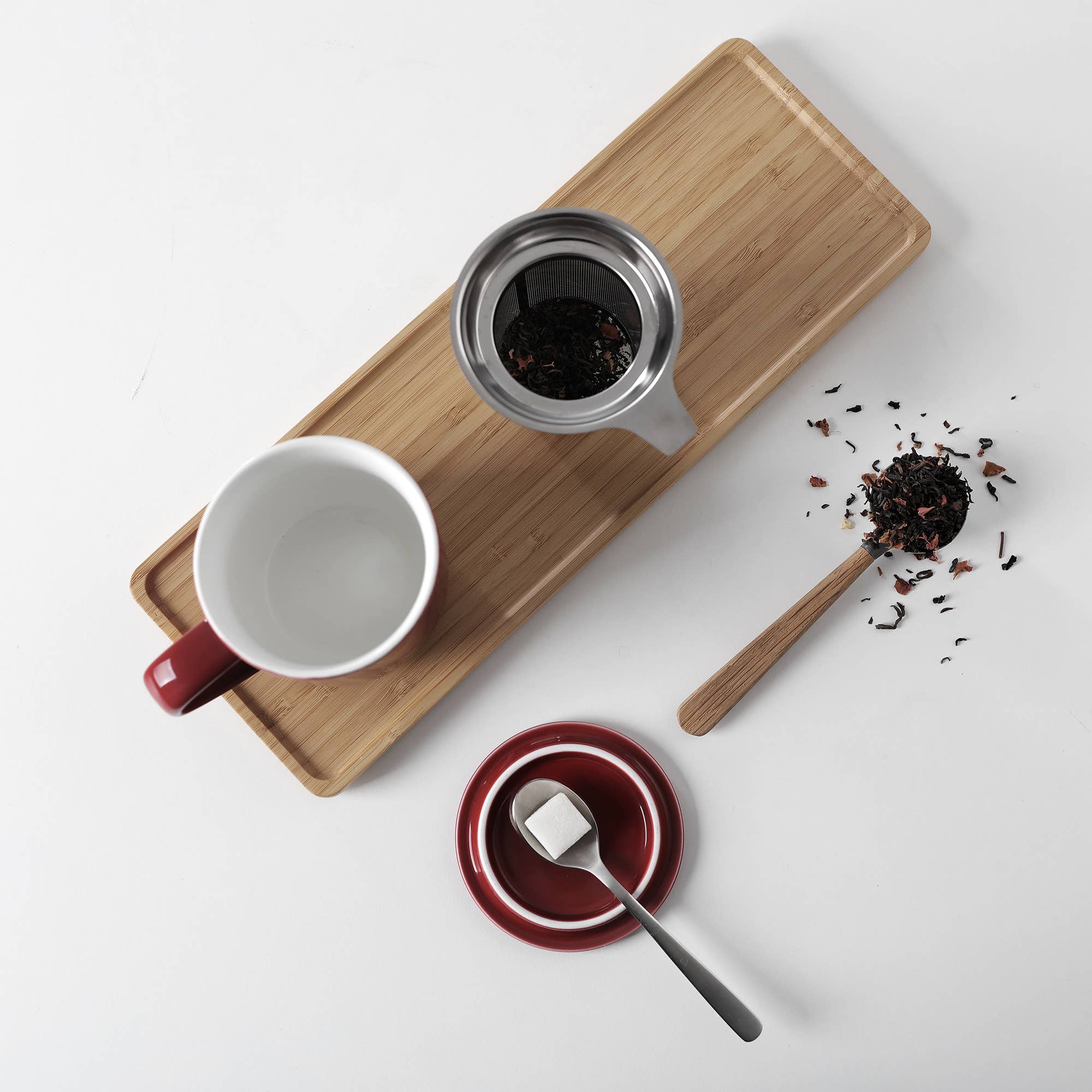 Tea-making setup with a white mug, wooden tray, tea leaves, and a spoon on a light gray background.
