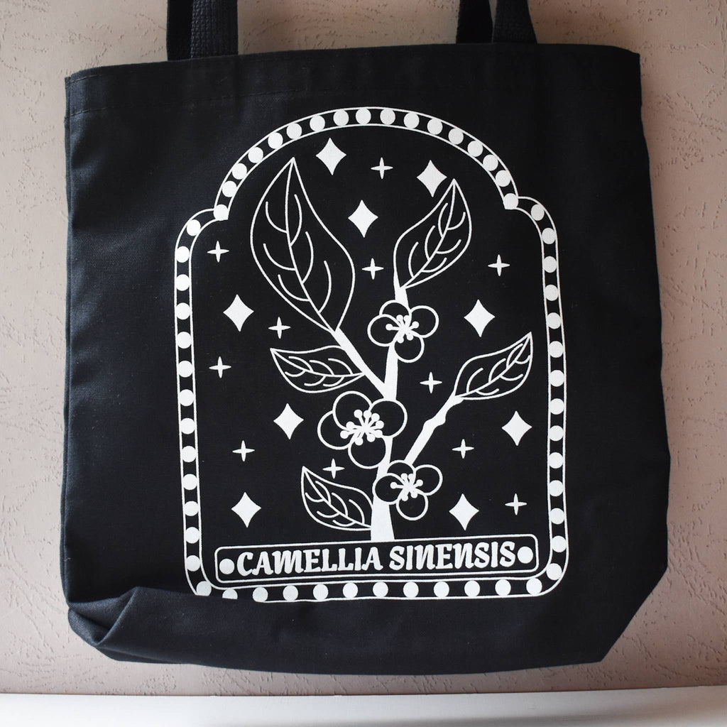 Black tote bag with white floral design and 'Camellia Sinensis' text on a neutral background
