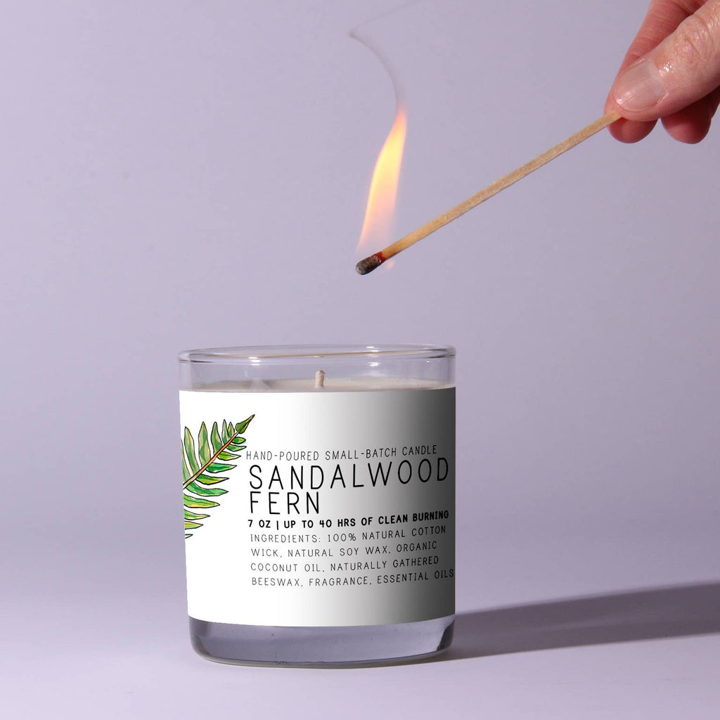 Candle labeled 'Sandalwood Fern' being lit with a match against a light purple background
