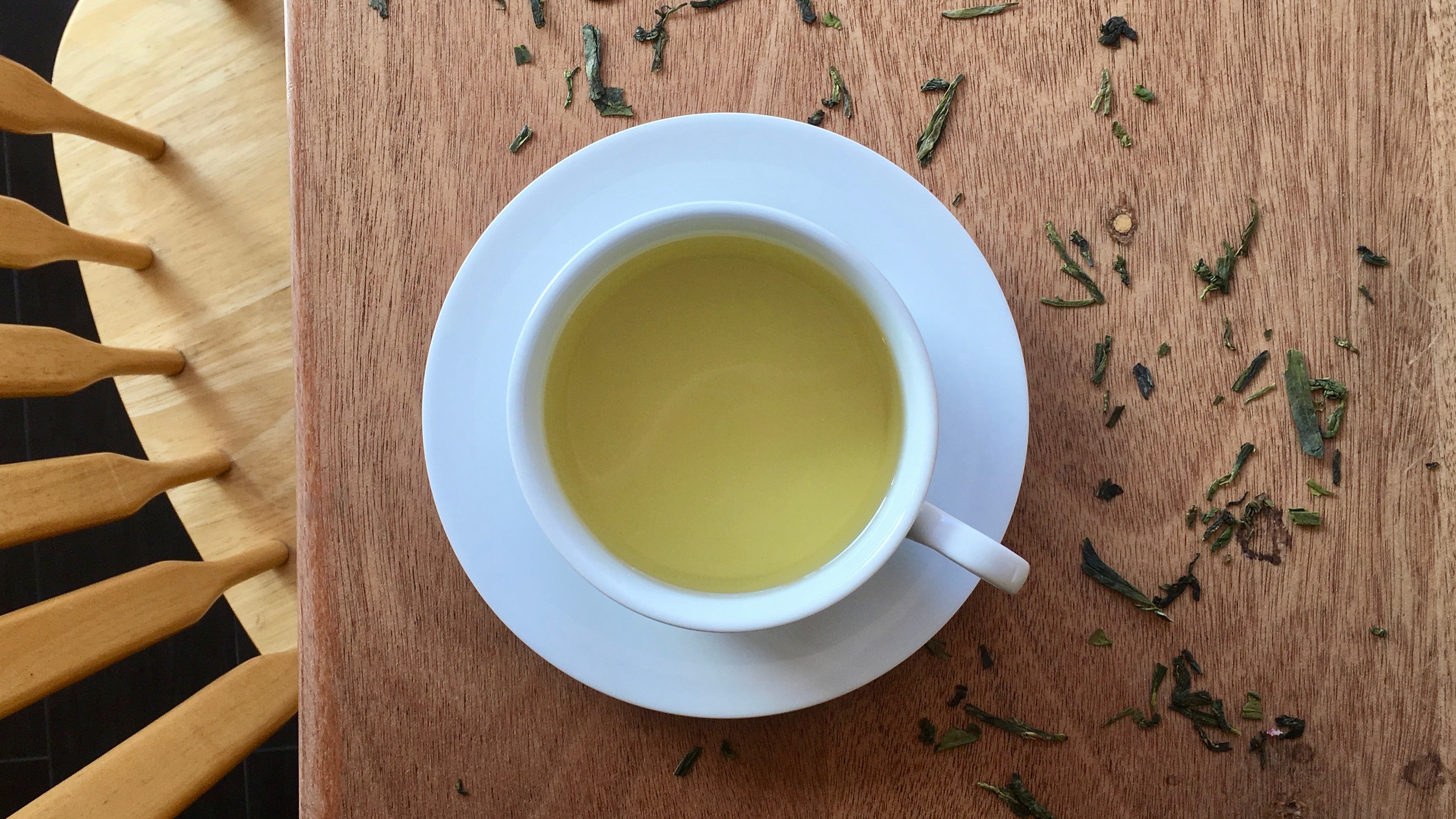 White cup of green tea on a wooden surface with tea leaves scattered around
