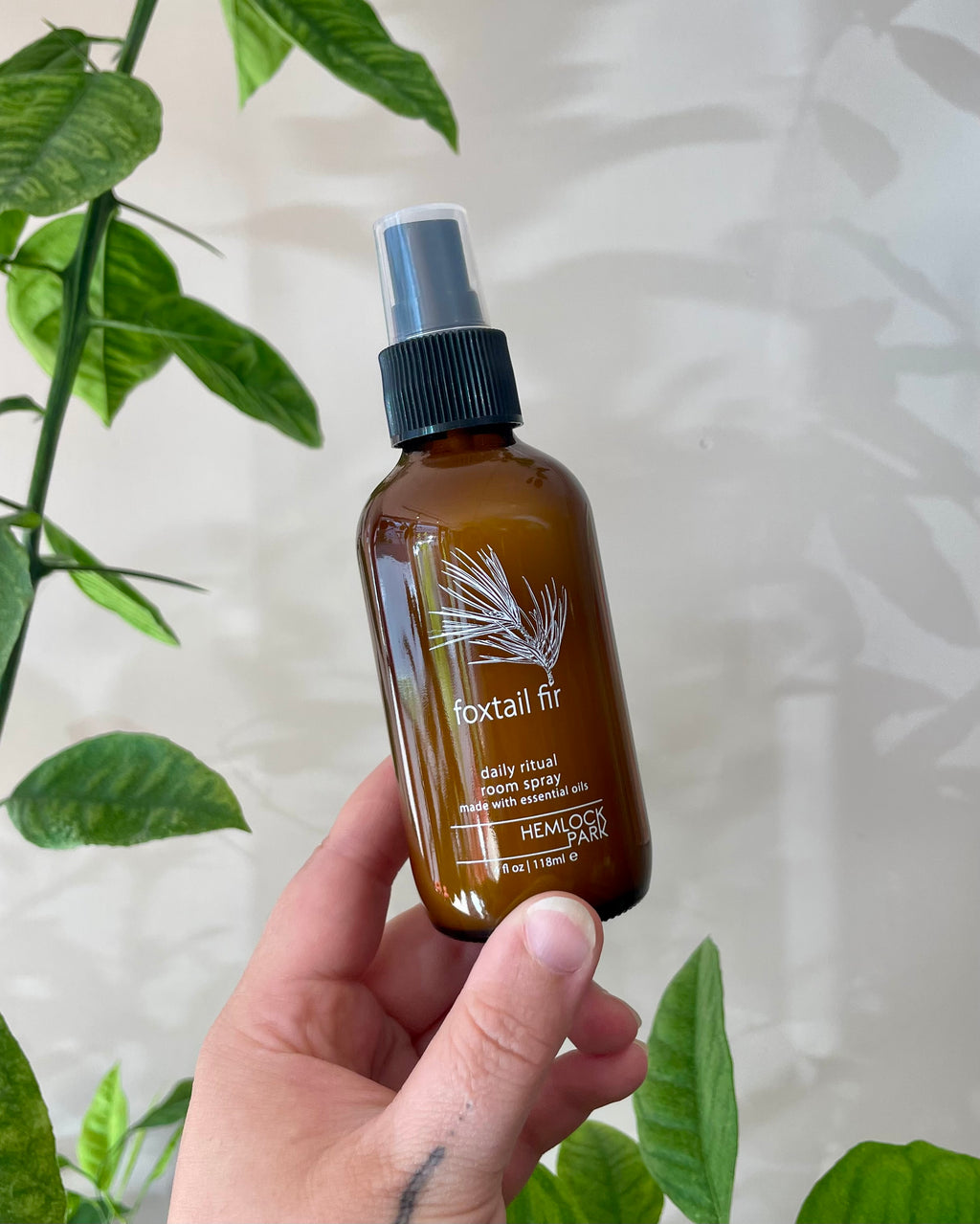 Hand holding a brown spray bottle labeled 'foxtail fir' with green leaves in the background