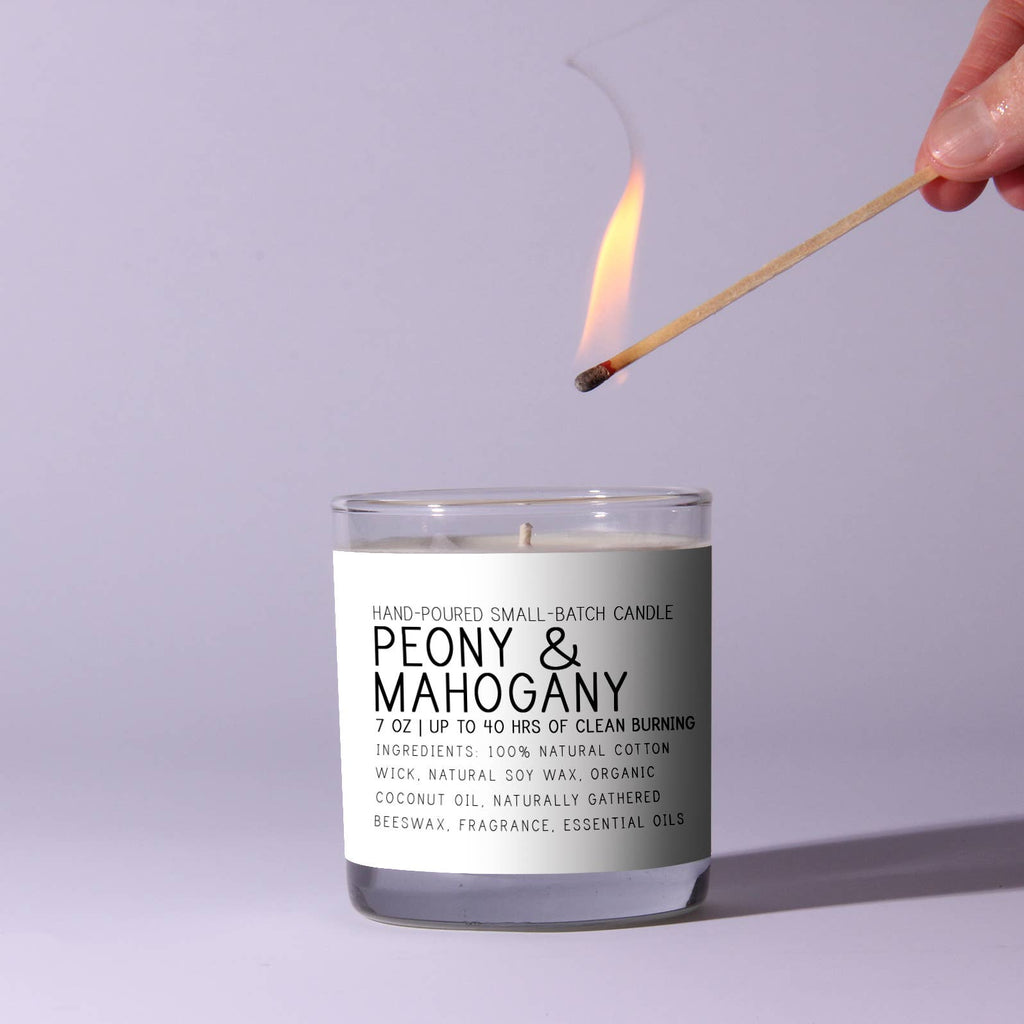 Candle labeled 'Peony & Mahogany' being lit with a match against a light purple background