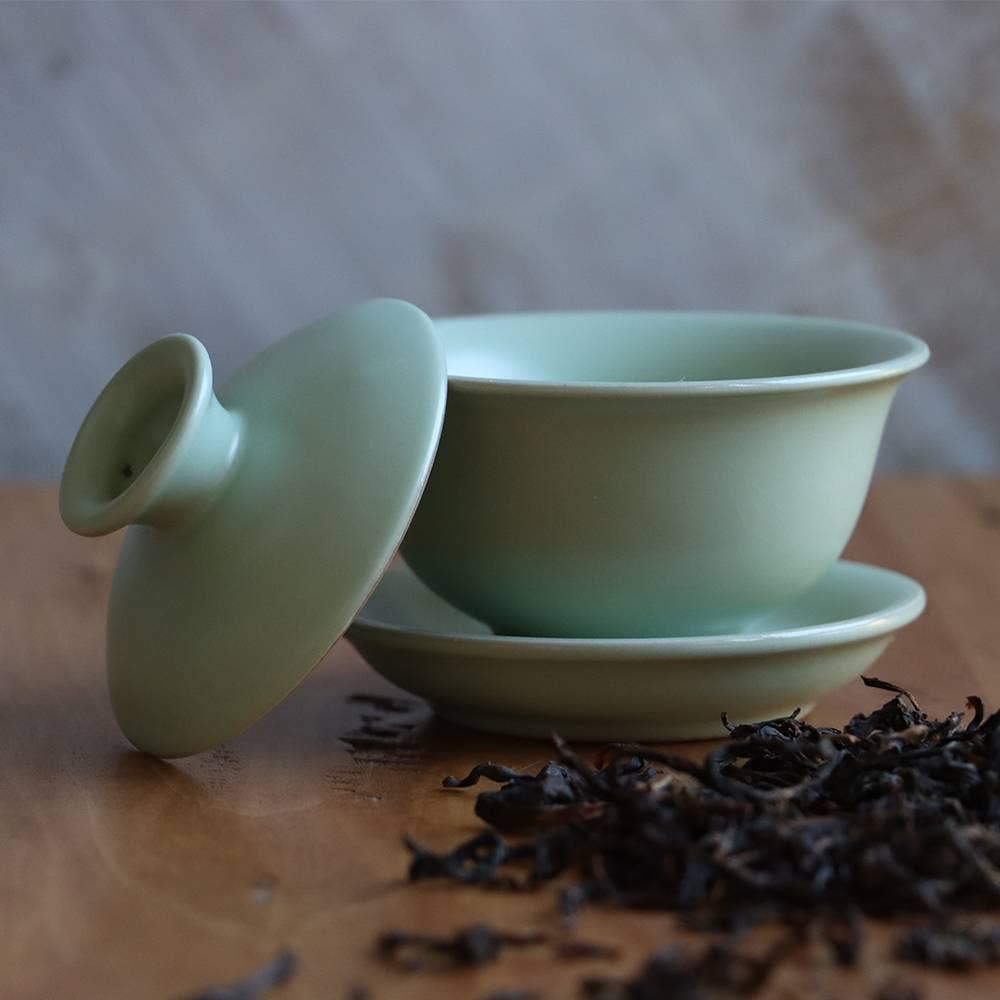 Light green ceramic teacup with lid on a wooden surface with tea leaves.