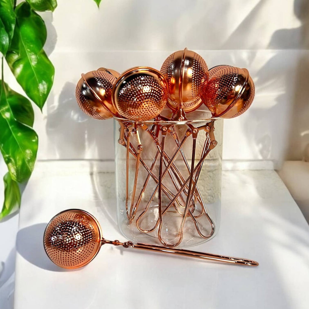 Set of rose gold tea infusers with a glass jar on a white surface.