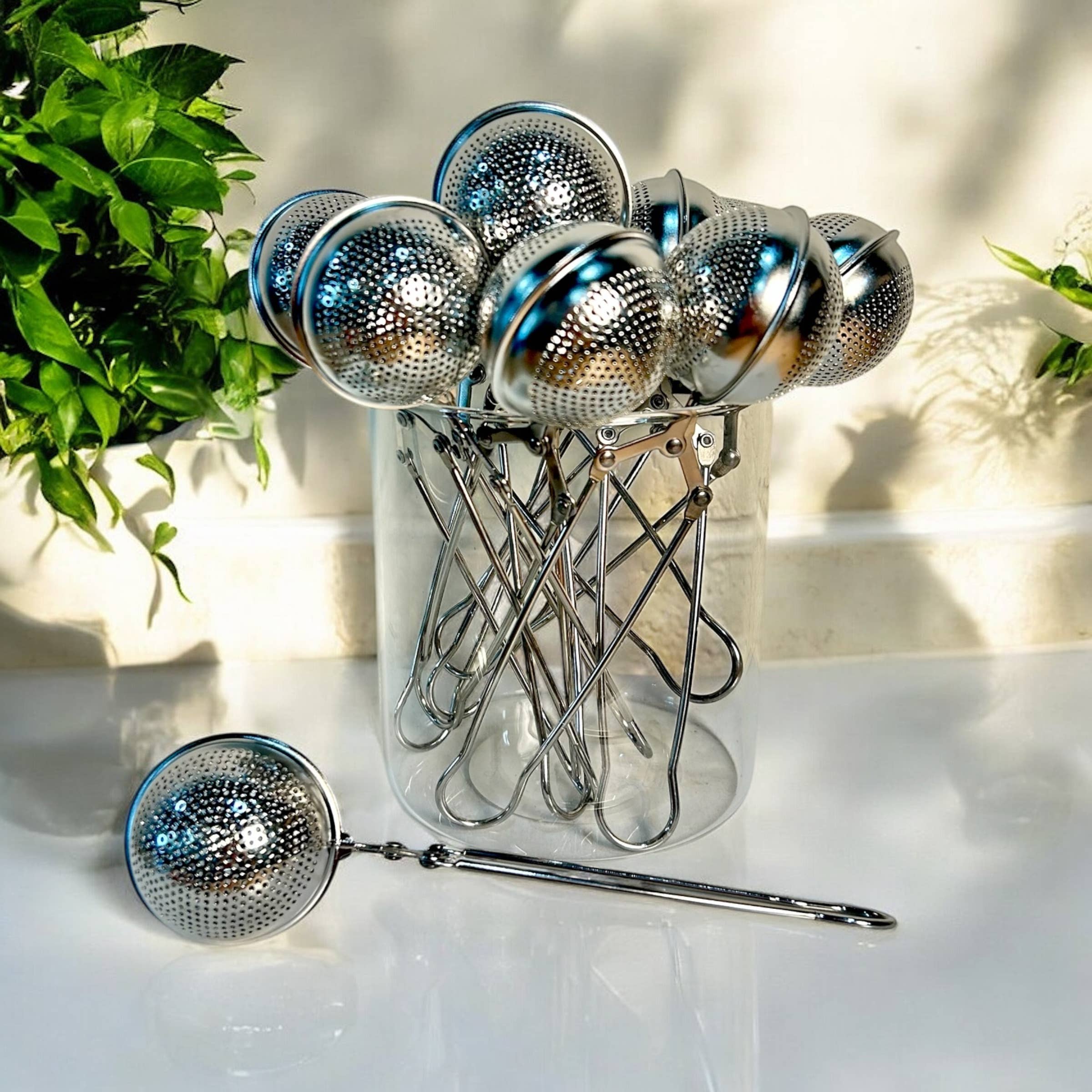 Set of metal tea infusers with a clear stand on a light surface with plants in the background