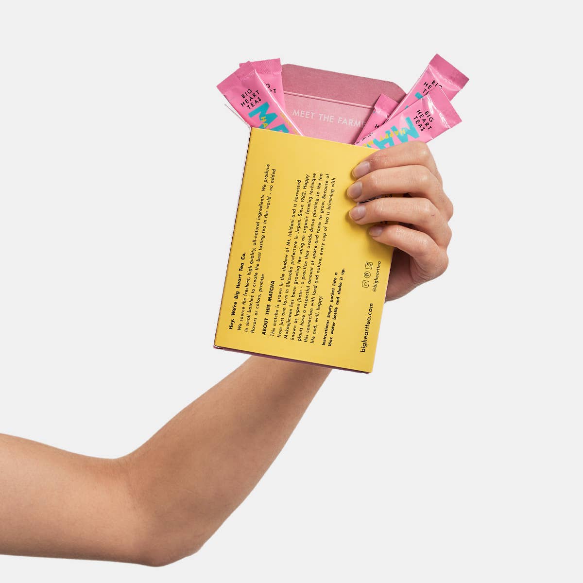 Hand holding a yellow box with pink packets of matcha on a light gray background