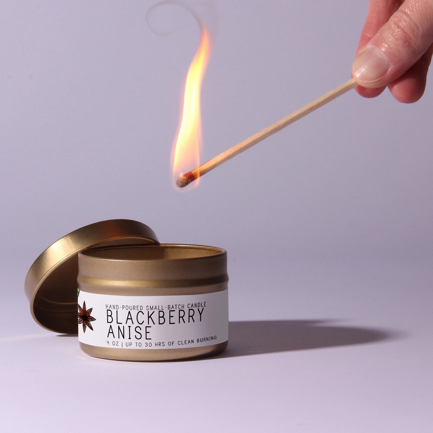Candle labeled 'Blackberry Anise' being lit with a match against a light purple background