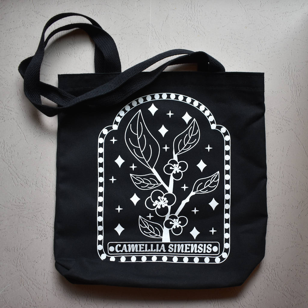 Black tote bag with white floral design and 'Camellia Sinensis' text on a gray surface