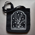 Black tote bag with white floral design and 'Camellia Sinensis' text on a gray surface