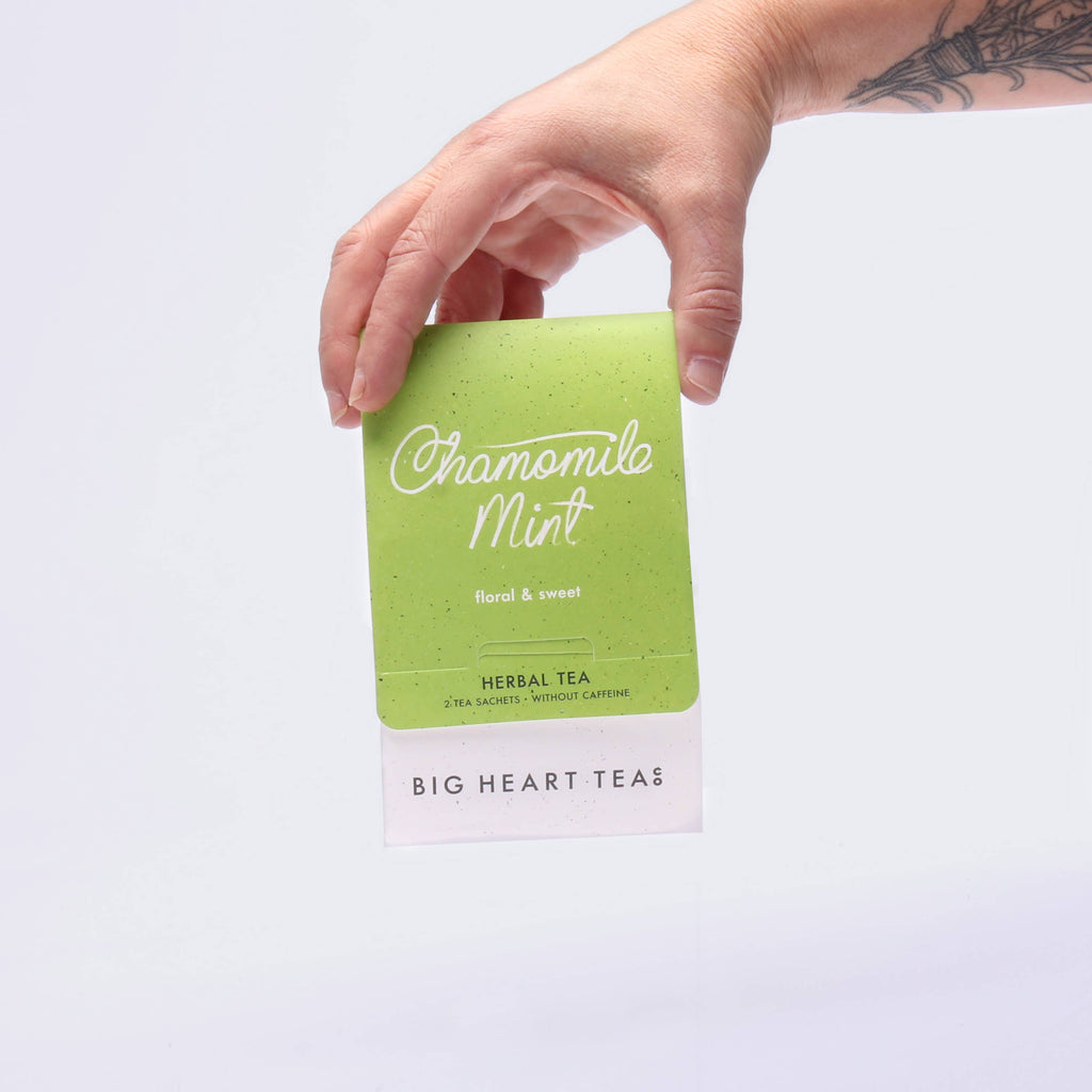 Hand holding a green tea package labeled 'Chamomile Mint' by Big Heart Teas on a light gray background