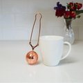 rose gold tea ball infuser next to a white mug on a light surface with flowers in the background.