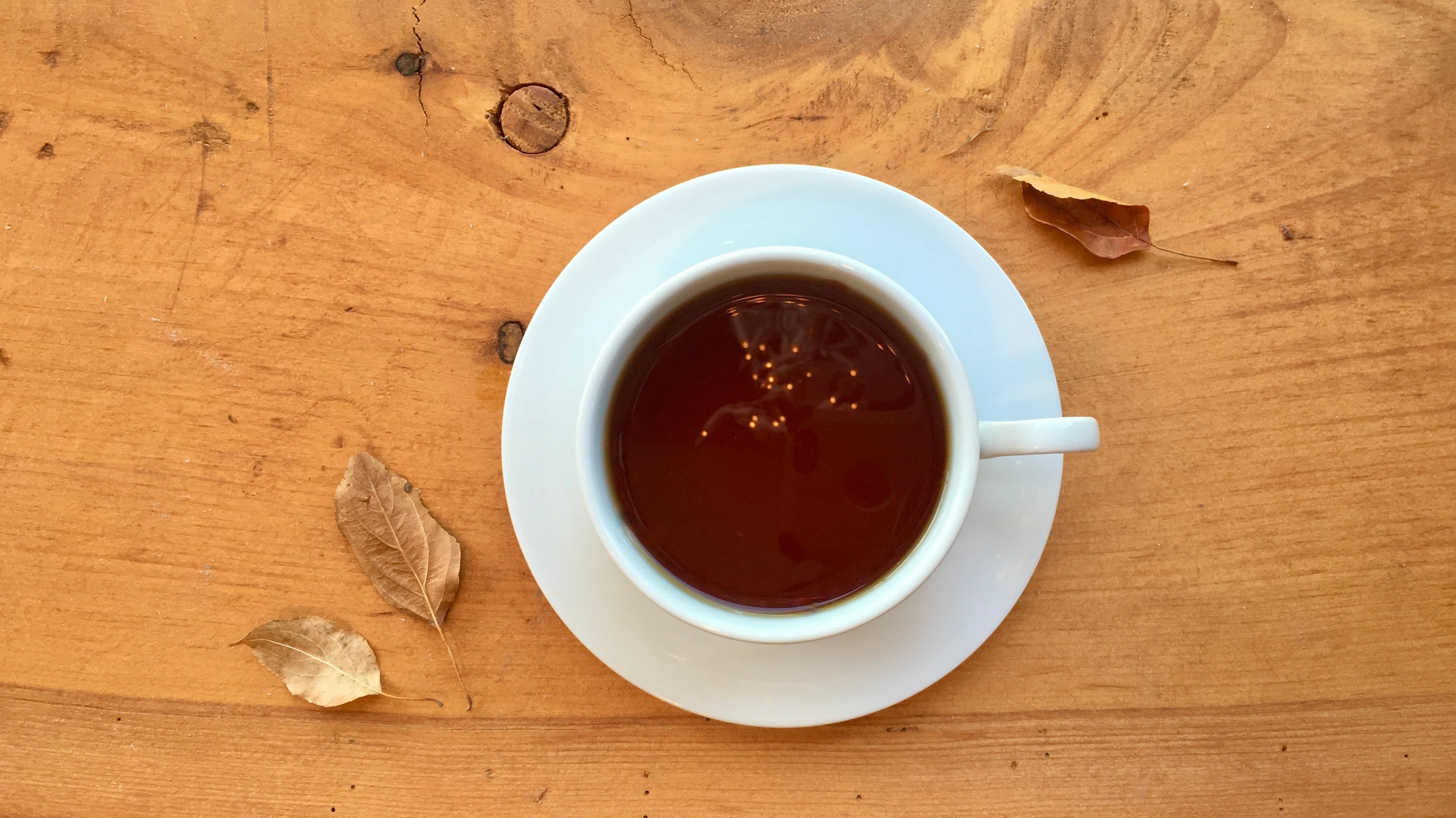 A white ceramic teacup sits on a matching saucer on a pale wooden table, filled with deep orange-brown tea reflecting tiny orange fairy lights. The table is sprinkled with yellow-brown fall leaves.