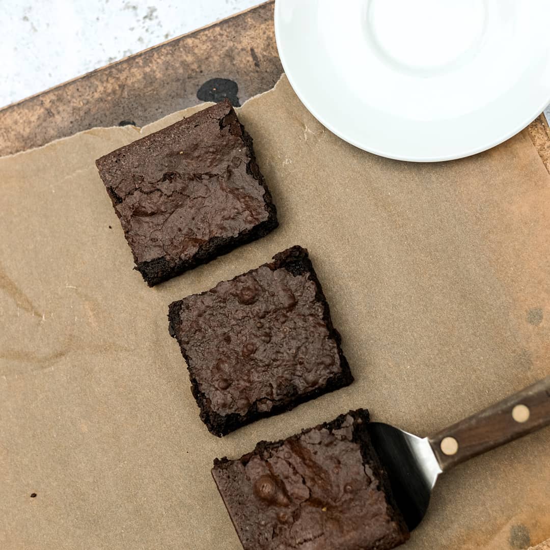 Three brownies on a piece of brown paper with a white plate and knife.